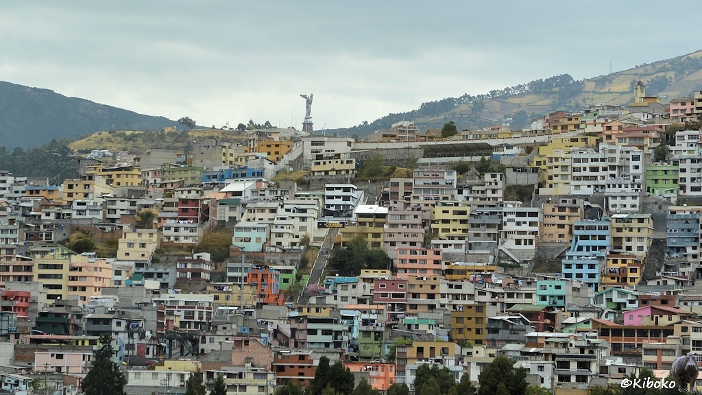 Das Bild zeigt zwei- und dreigeschossige, bunte Häuser an einem Berghang. Eine Treppe führt zwischen den Häusern nach oben. Oben auf dem Berg steht ein großes, silbernes Denkmal.
