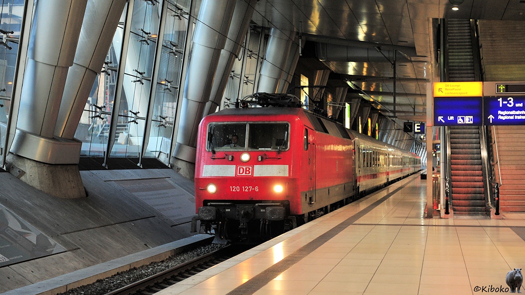 Das Bild zeigt eine rote Elektrolokomotive mit einen weißen Balken auf der Front. Die Lok zieht 6 weiße Personenwagen, die einen roten Streifen unterhalb der Fenster haben. Der Zug fährt an einen Bahnsteig aus hellen Platten ein. Der Zug ist in einer Bahnhofshalle mit silbernen runden Säulen und Glasfronten. Am rechten Bildrand führt eine Treppe nach oben.