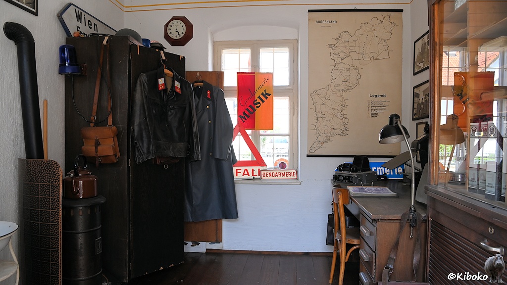 Das Bild zeigt die Innenansicht eines Polizeipostens. Rechts steht ein Schreibtisch mit Lampe. Rechts steht ein eiserner Holzofen und ein Schrank mit Polizeimänteln. Im Hintergrund ist eine Karte vom Burgenland.
