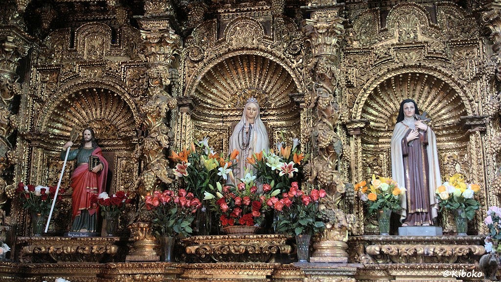 Das Bild zeigt drei Nischen im Seitenbereich. In der Mittleren Nische steht eine Marienstatue. Sie ist flankiert von zwei weiteren Figuren. Die Nische ist mit roten Rosen und anderen Blumen reichlich dekoriert. Zwisschen den Nischen sind Säulen mit Weinreben und Weintrauben schnitzerein. Alle ist reichlich verziert und vergoldet.