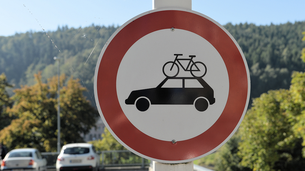 Das Bild zeigt ein rundes Schild mit rotem Rand. Darauf ist ein schwarzes Auto auf dem ein Fahrrad auf dem Dach steht auf weißem Grund.