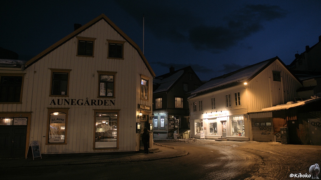 Das Bild zeigt eine Straßenecke mit weißen Holzhäusern. Die Schaufenster im Ergeschloss sind beleuchtet. Der Himmel ist blau.