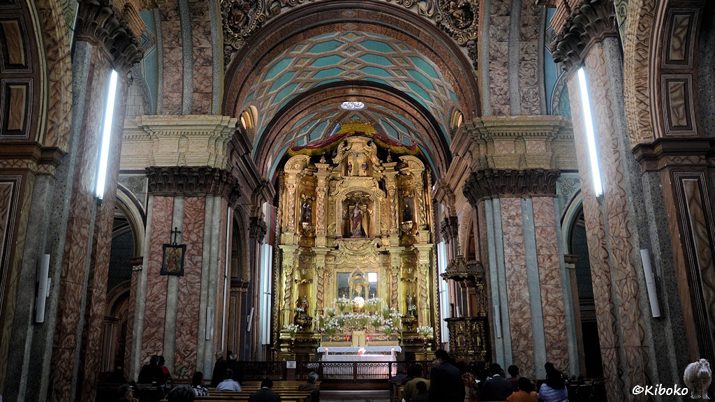 Das Bild zeigt einen Innenansicht einer Kirche. Massive Bögen sind mit rötlich gemusterten Steinen dekoriert. An den Kanten und in der Mitte sind grau-blaue Steinplatten. Zentral ist der golden Hochaltar, der über mehrere Etagen geht. Alle Schnitzereien und Verzierungen sind vergoldet. Am unterren Bildrand sitzen Leute und lauschen der Messe.