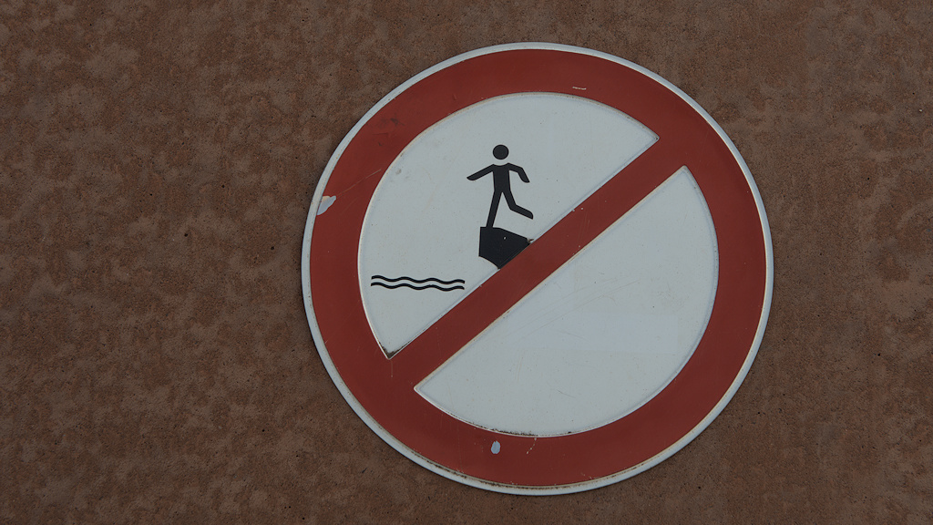 Das Bild zeigt ein rundes Schild mit retem Rand. Darauf ist eine schwarze Figur, die auf einem Bein auf einem Vorsprung steht. Darunter sind Wellenlinien. Ein roter diagonler Streifen geht von links unten nach rechts oben durch das Schild.