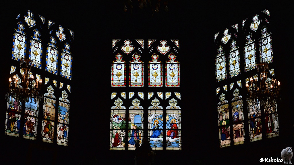 Das Bild zeigt drei bunte Kirchenfenster hinter dem Altar einer Holzkirche. An den Seiten hängt jeweils ein Kronenleuchter vor den Fenstern.