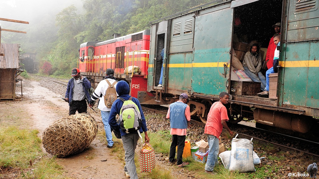Das Bild zeigt den grünen Gepäckwagen mit offener Tür und die rote Diesslok von hinten. Mehrere Menschen sitzen im Gepäckwagen auf der Ladung. Ein paar Säcke, Kartons und leere Körbe liegen vor der offenen Tür des Gepäckwagens. Ein Mann trägt eine Kutte mit der Aufschrift: Securité FCE und führt Protokoll.