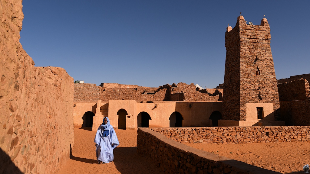 Das Bild zeigt den Innenhoh einer Moschee mit einem sandigen Weg. Hinten ist wieder das verputzte Gebäude mit den pilzförmigen Eingängen. Am rechten Bildrand steht der viereckige Turm. Ein Mann im langen hellblauen Gewandt verlässt die Moschee.