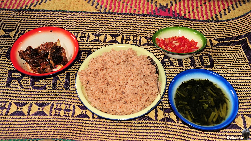 Das Bild zeigt viert gut gefüllte Schalen auf einer Schilfmatte. In der Mitte ist eine größere Schale mit Reis. Links ist eine kleiner Schale mit Fleisch und Knochen, rechts sind zwei Schalen mit Tomatenstücken und eingelegtem Grünzeug.