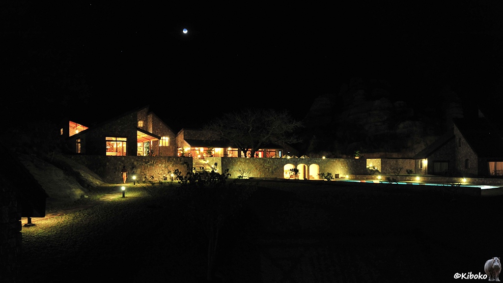 Das Bild zeigt ein verwinkeltes, hell erleuchtetes Gebäude. Der Weg und die Außenmauern sind durch Lampen ausgeleuchtet. Am rechten Bildrand ist das blau leuchtende Schwimmbecken. Darüber ist tiefschwarze Nacht mit einem Scihelförmigen Mond über dem Gebäude.