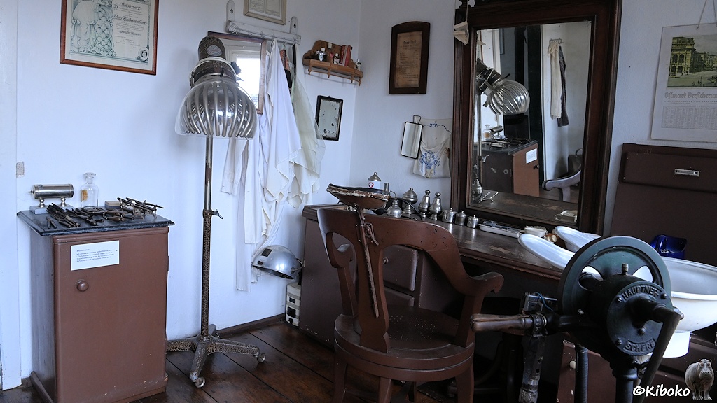 Das Bild zeigt einen historischen Frisörsalon mit Holzstuhl, Spiegel, Waschbecken und Trockenhaube. Am linken Bildrand liegen eiserne Lockendreher die erhitzt werden müssen.