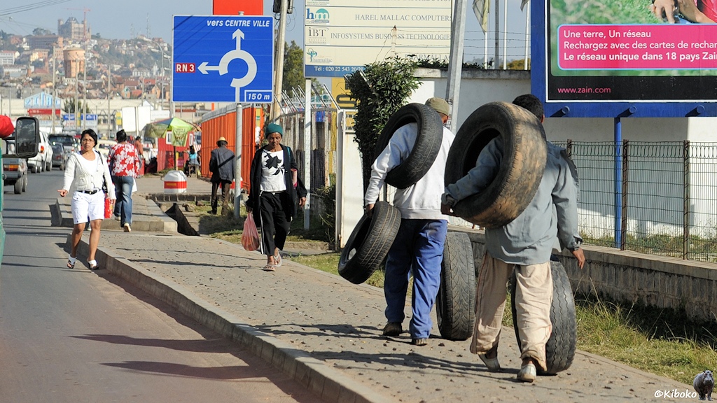 Zwei Männer tragen und rollen 5 Autoreifen in unterschiedlichen Größen auf dem Geheweg.