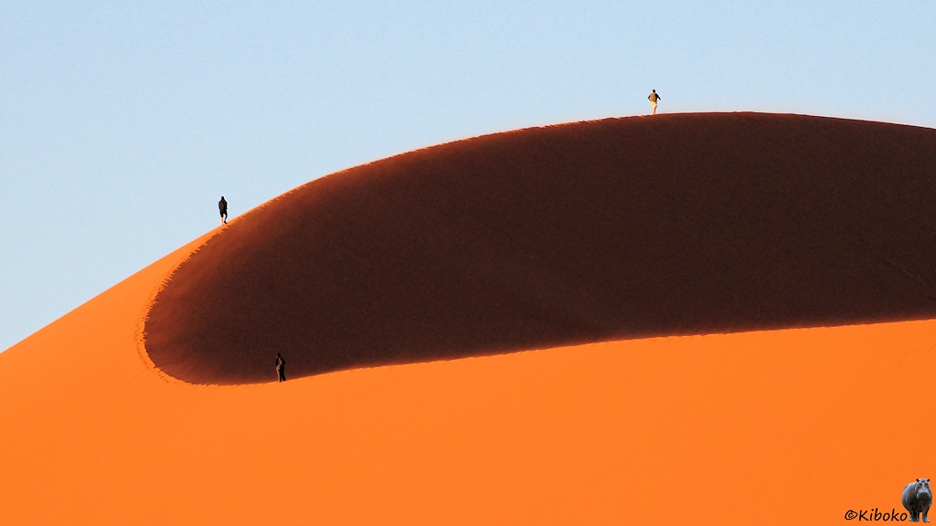 Drei Menschen laufen auf dem Bogen eines Dünenkammes. Der Wind fegt feinen Sand über den Kamm.