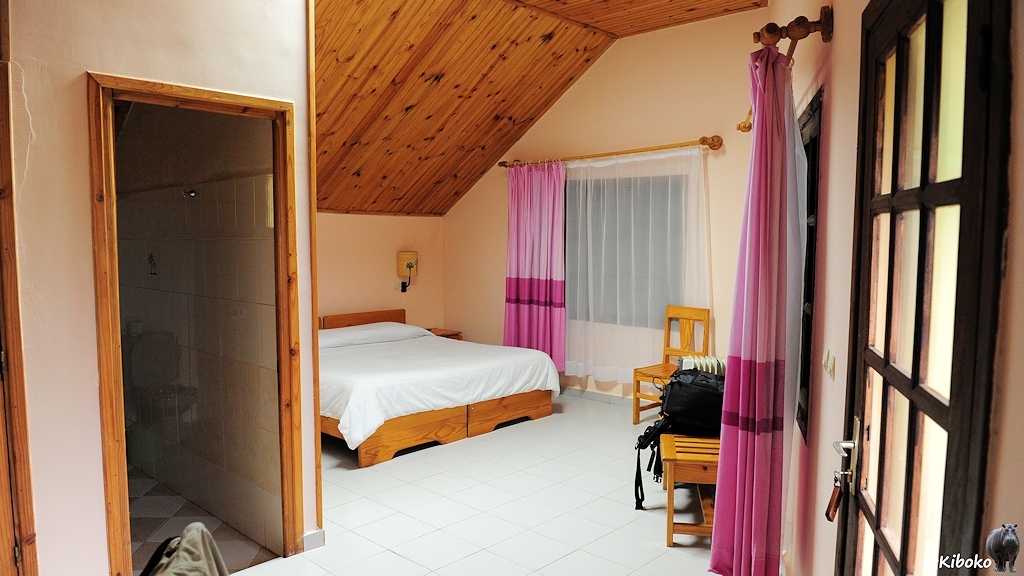 Das Bild zeigt die Innenansicht vom Hotelzimmer mit beigen Wänden, hellen Bodenflißen. Mögel und Decken sind aus hellem Kiefernholz. Das Bett wird hinter einer Ecke mit dem Bad leicht verdeckt.