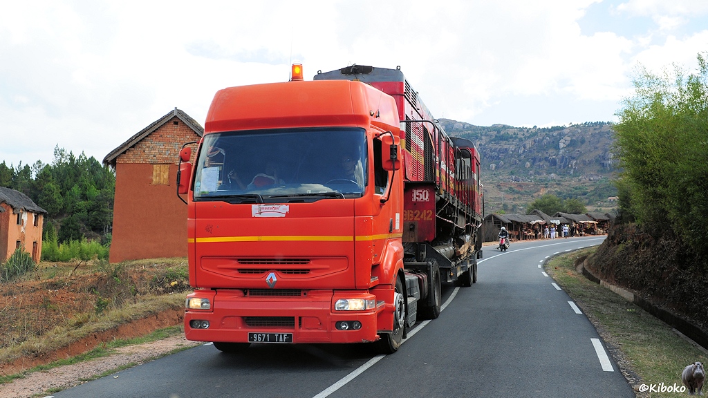 Ein orangeroter, moderner Renault-LKW mit Warnleuchte und Tiefladeranhänger kommt auf einer schmalen Teerstraße entgegen. Auf dem Auflieger steht eine rote Diesellok ohne Räder.