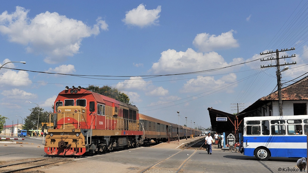 Das Bild zeigt eine orangerote-beige Diesellok mit einen Schnellzug auf einem Bahnübergang. Die Wagen stehen an einem Bahnsteig. Am rechten Bildrand drängelt sich ein weiß-blauer Bus in das Bild.