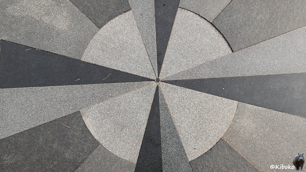Das Bild zeigt eine Fläche aus grauen Granitsteinen. In der Mitte ist ein kleiner kreisförmiger Stein aus dunklem Granit. Davon geht jeweils ein paar spitzwinkliger Dreiecke in jede der vier Himmelsrichtungen. Die Flächen dazwischen füllen hellgrauer Granit zu einem Kreis.