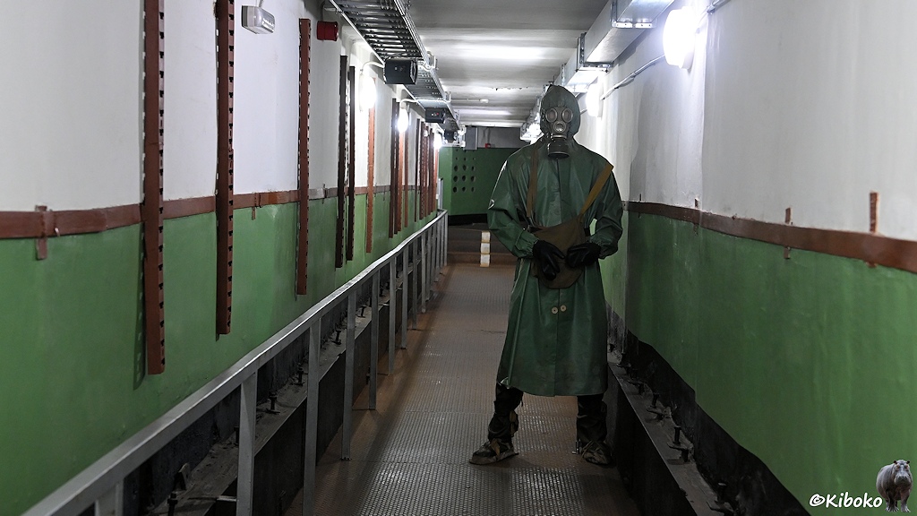 Das Bild zeigt einen langen Gang. Die Wände sind oben weiß und unten grün lackiert. Der Boden besteht aus rostigem Riffelblech. Im Gang steht eine Puppe mit Gasmaske und ABC-Schutzbekleidung.