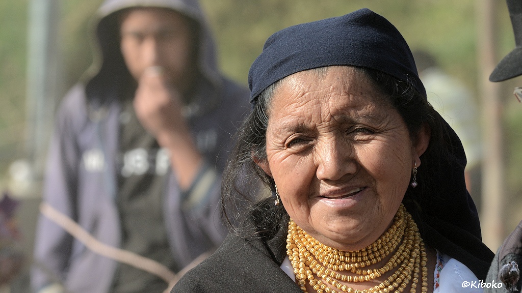 Das Bild zeigt das Porträt einer älteren Frau mit dunkelblauem Kopftuch, schwarzem Umhang, weißer Bluse und 11-fach um den Hals gewickelter Goldkette.