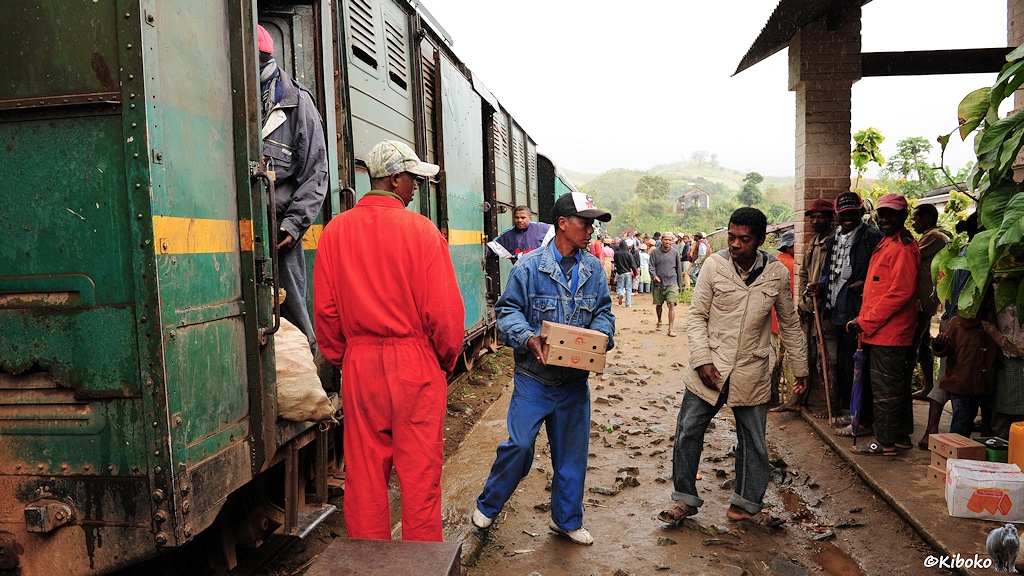 Das Bild zeigt den Gepäckwagen mit offener Tür. Männer haben eine Kette gebildet. Ein Papkarton wird zum überdachten Bahnhof durchgereicht. In der Tür schaut ein Sack heraus.