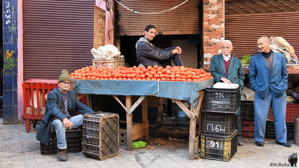 Das Bild zeigt einen Laden mit halgeöffnetem Rolltor. Davor steht ein einfacher zusammengezimmerter Holztisch mit Tomaten. Danebne stehen schwarze Plastikstiegen. Ein Mann sitzt auf einer Stiege vor dem Tisch. Zwei weitere Männer stehen daneben. Einer steht hinter dem Tisch.