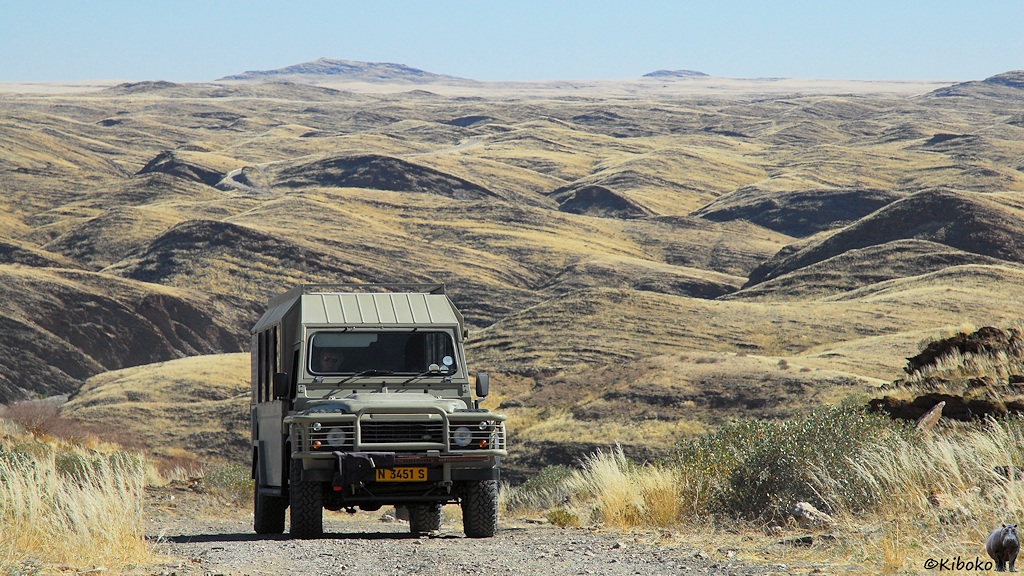 Ein beigegrüner Landrover fährt auf einerm Schotterweg über einen Pass mit grasbewachsenem Hügeln im Hintergrund.