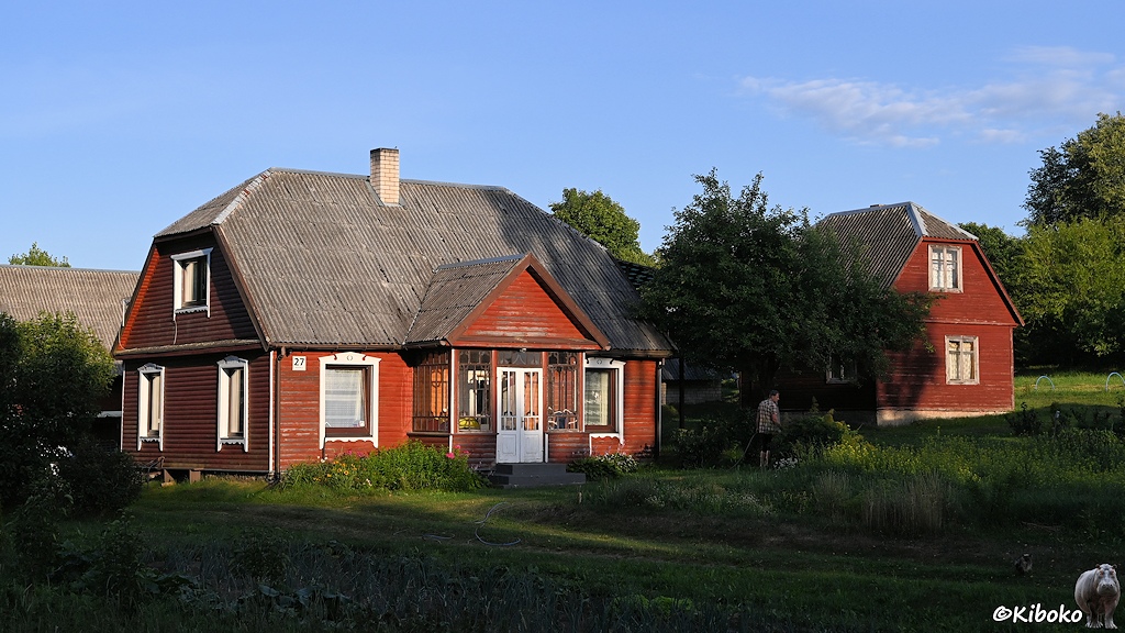 Das Bild zeigt ein rotbraun gestrichenes Holzhaus mit Krüppelwalmdach aus gewellten Zementplatten. In der Mitte ist ein Anbau mit dem Eingangsbereich. Rechs steht ein Nebengebäude in ähnlicher Bauart.