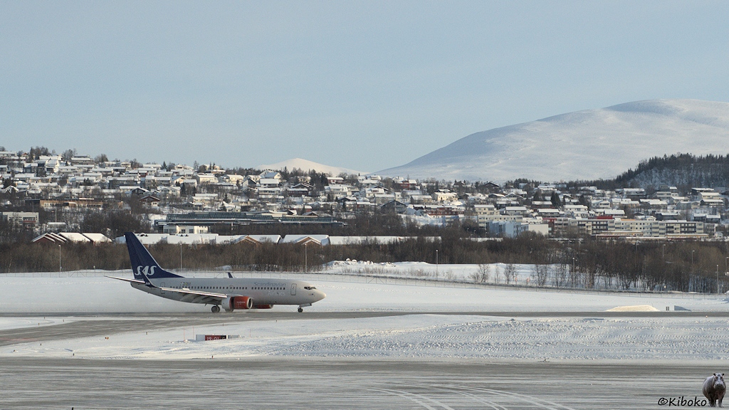 Das Bild zeigt einen kleinen weißen zweistrahligen Düsenflieger mit blauen Leitwerk und roten Turbinen auf der Landebahn eines verschneiten Flughafens. Im Hintergrund stehen Häuser mit schneebedeckten Dächern.