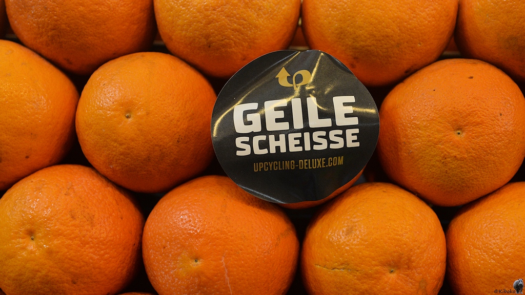 Nahaufnahme von orangen an einem Verkaufsstand. Eine Orange hat einen großen schwarzen Aufkleber mit der Aufschrift: Geile Scheisse.