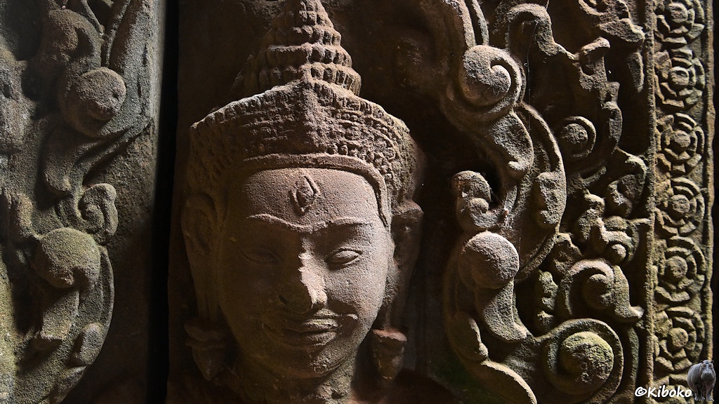 Das Bild zeigt einen Kopf mit abgebrochener Nase, verzierten Haaren oder Kopfbedeckung und rautenfömigen Amulette auf der Stirn als Relief. Darum sind reiche Verzerierungen in den Sandstein gehauen.