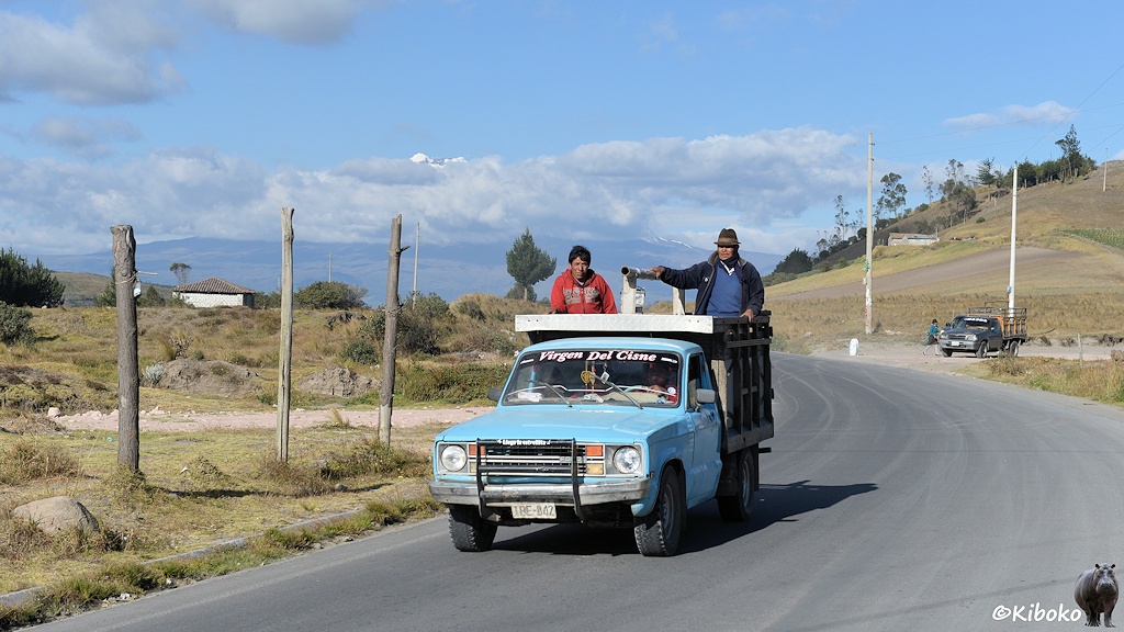 Das Bild zeigt eine geteerte Bergstraße. Ein hellblauer Ford Pick-up kommt entgegeben. Auf der hochbordigen Ladefläche stehen zwei Männer. Auf der Windschutzscheibe steht in weißen Buchstaben: Virgen Del Cisne. Im Hintergrund sit ein schneebedeckter Vulkan. Nur die Spitze schaut aus den Wolken heraus.