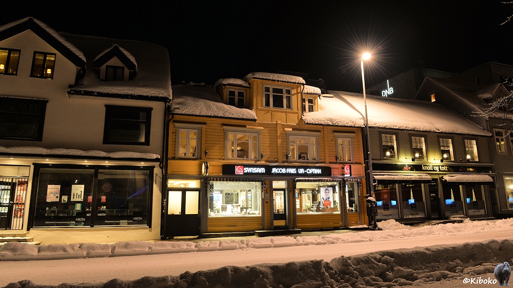 Das Bild zeigt ein zweigeschossiges Holzhaus mit beleuchteten Schaufenster im Erdgeschoss. Neben dem Erker sind Schneeberge auf dem Dach.