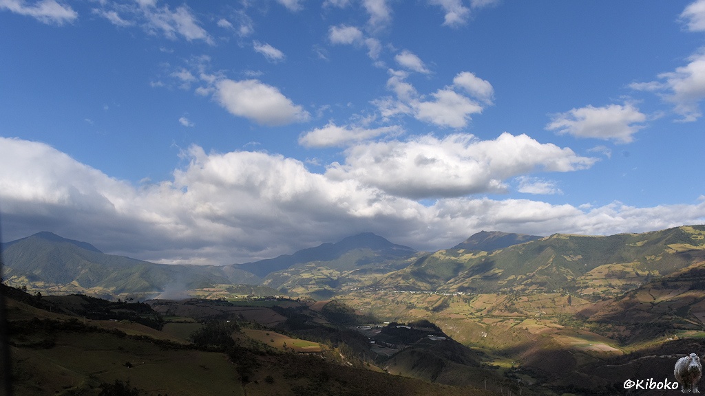 Das Bild zeigt eine Landschaft mit einem weiten Tal mit bewaldeten Bergen im Hintergrund. Über dem Tal ziehen einzelne Wolken, die ihren Schatten ins Tal werfen.