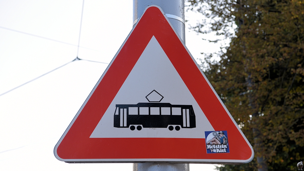 Das Bild zeigt ein dreieckiges Schild mit der Spitze nach oben mit rotem Rand. Darauf ist ein schwarzer vierachsiger Straßenbahnwagen mit Serenstromabnehmer auf weißem Grund. An der unteren rechten Ecke ist ein Aufkleber mit dem Text: Holstein Kiel.