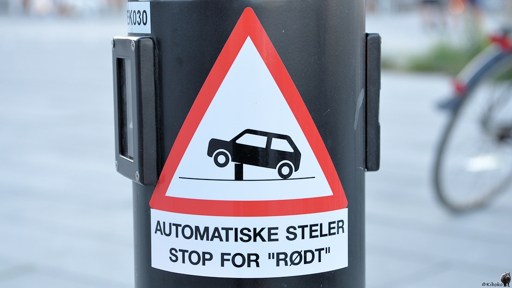 Das Bild zeigt ein dreieckiges Schild mit der Spitze nach oben und rotem Rand. Darauf ist ein schwarzes Auto auf weißem Grund. Der Vorderwagen ist angehoben und unter der Vorderachse ist ein Klotz. Darunter ist ein rechteckiges Schild mit dem Text: Automatiske Steler Stop for Rodt.