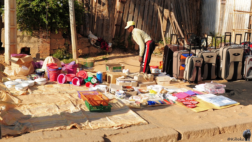Das Bild zeigt einen Verkaufsstand auf dem Geheweg. Ein Mann stellt auf Matten weüße Kochtöpfe und rosafarbene Platikeimer auf. Daneben stehen ein gutes Dutzen Rollkoffer in unterschiedlichen Größen.