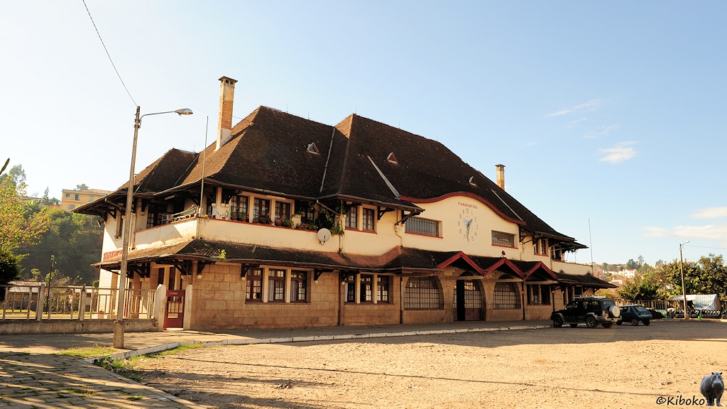 Das Bild zeigt ein zweigeschossiges Gebäude mit einem dunklen ineinandergeschachtelten Walmdach. Das Erdgeschoss besteht aus hellem Sandstein. Das erste Stock ist hellbeige angemalt. In der Mitte des ersten Stocks ist eine große Wanduhr. Vor dem Gebäude parken auf einem Schotterplatz zwei Autos .