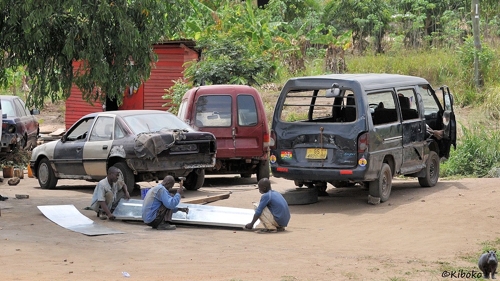 Drei Männer hocken auf dem staubigen Boden und bearbeiten ein großes Blech. Im Hintergrund stehen verbeulte Autos.