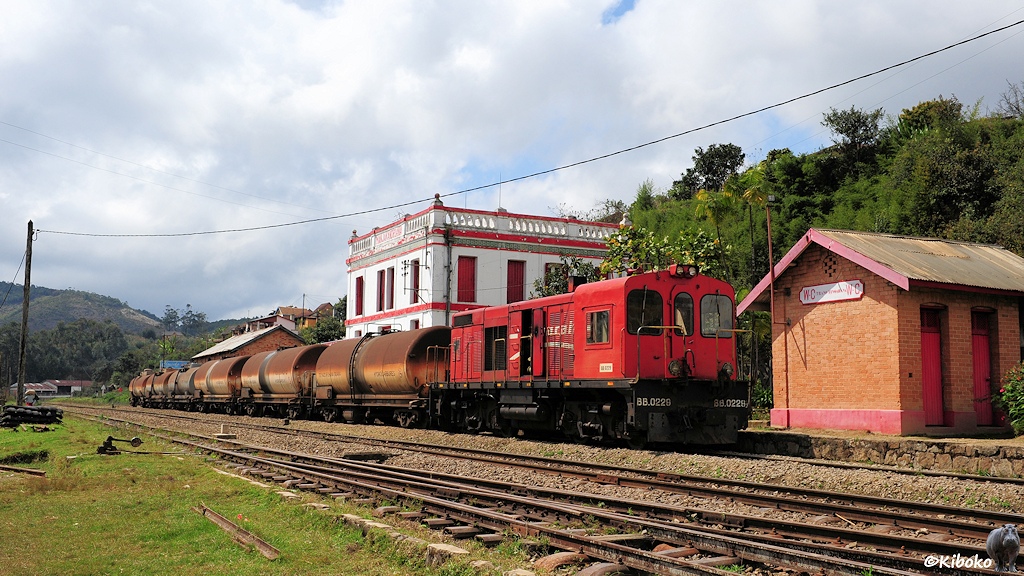 Eine rote, vierachsige Diesellok mit breitem Führerhaus steht mit sechs verdreckten Kesselwagen in einem dreigleisigen Bahnhof. Im Hintergrund ist ein weißes, würfelförmiges Bahnhofsgebäude mit roten Fensterläden und Zierstreifen.