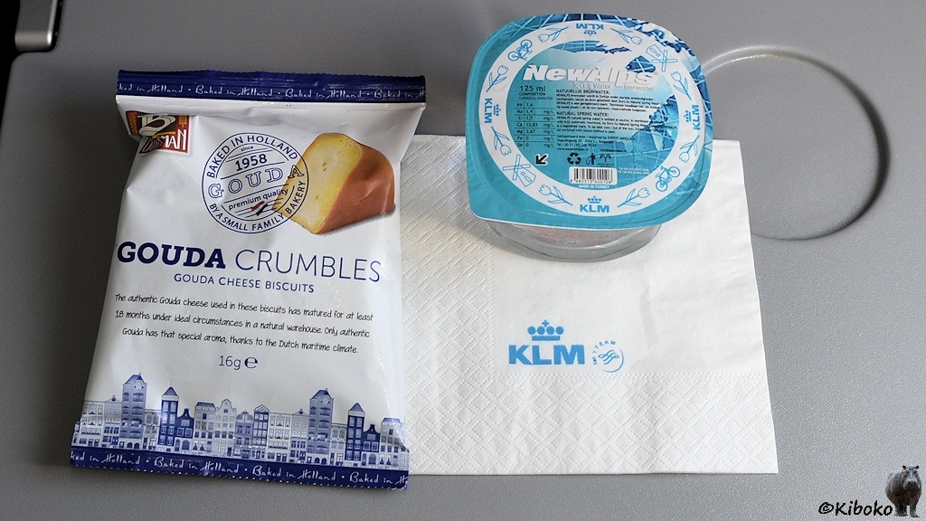 Das Bild zeigt das heruntergeklappte Tischen. Darauf ist eine Serviette mit der Aufschrift KLM. Ein Plastikbecher mit hellblauem Decke mit der Aufschrift NewAlps. Der enthält Wasser. Links daneben liegt eine kleine weiß-blaue Plastiktüte mit der Aufschrift: GOUDA Crumbles.