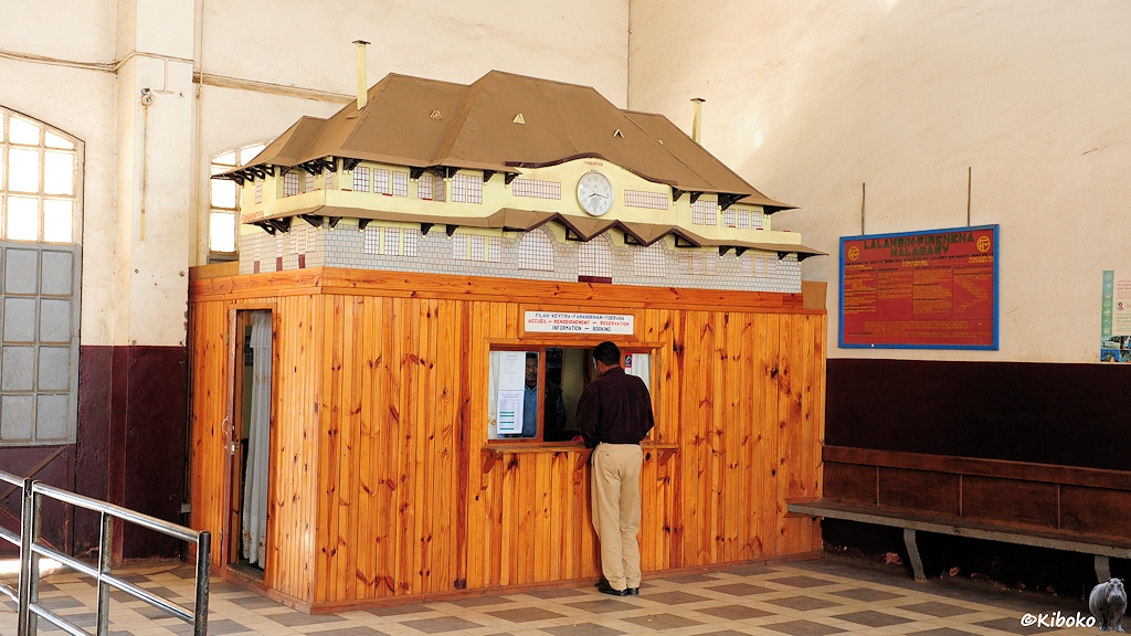 Das Bild zeigt einen hölzernen Schlater innerhalb eines Warteraumes. Vor dem geöffneten Schalterfenster steht ein Mann. Auf dem Dach des Fahrkartenschalters ist ein Modell des Bahnhofs aufgebaut. Die Wartezimmeruhr ist am Modell angebaut.