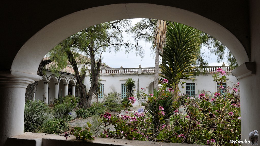 Das Bild zeigt den Blick durch einen Rundbogen in den Garten. Der Garten ist durch eingeschossige, weiße Gebäude begrenzt. Die Wege sind gepflastert. Es gibt viele Blumen mit rosa Blüten, ein paar hohe Bäume und ein paar kleine Palmen.