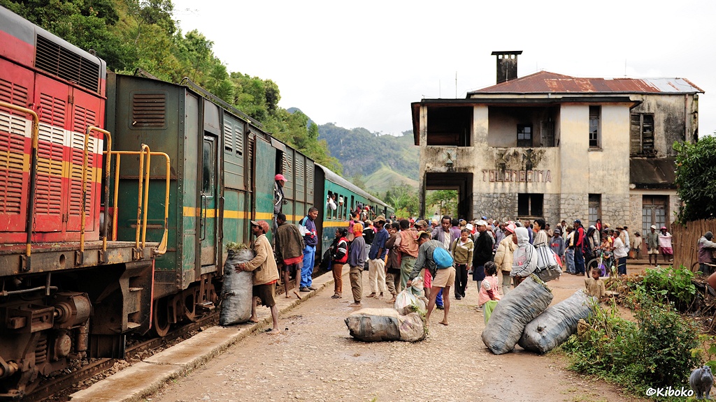 Das Bild zeigt den Gepäckwagen und weitere Wagen vom Zug. Im Vordergrund sind große graue Säcke auf dem Bahnsteig. Dahinter sind viele Menschen in bunten Kleidern. Im Hintergrund ist das zweistöckige Bahnhofsgebäude. Die Wände sind vom Klima schwarz verwittert.