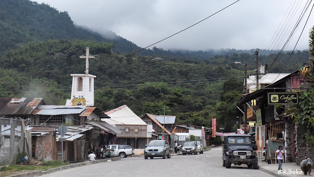 Das Bild zeigt eine gepflasterte Straße in einem Ort. Autos stehen am Straßenrand. Ein kleiner eckiger Krichturm mit einen Kreuz auf dem Flachdach überragt die Häuser. Im Hintergrund sind bewaldete Berge. Wolken ziehen über die Berge.