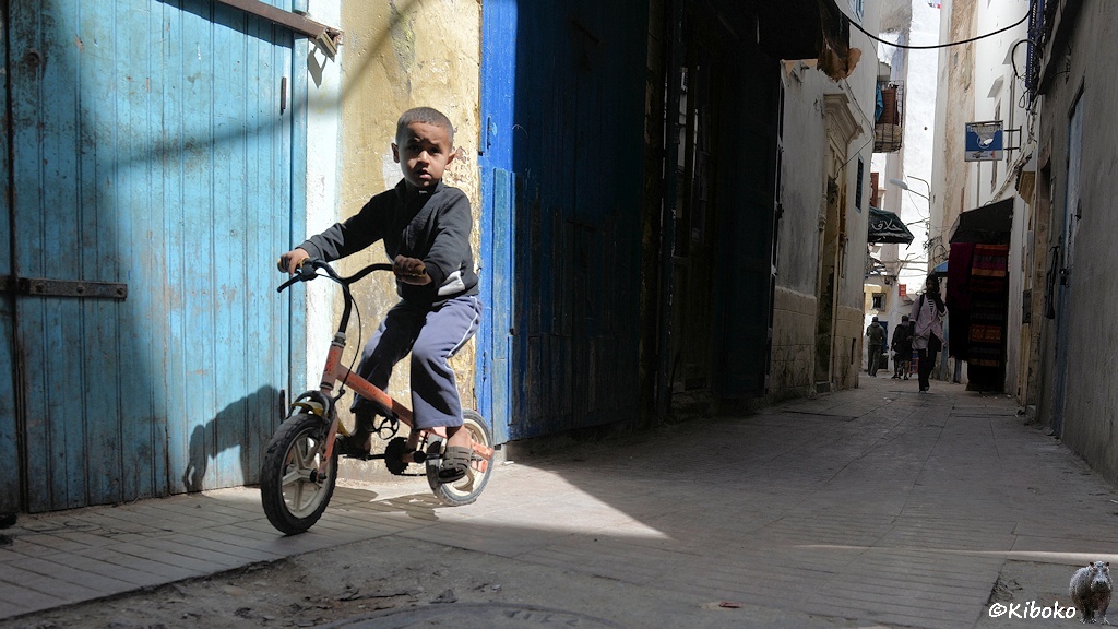 Das Bild zeigt eine Gasse im Schatten. Die Sonne bescheint ein Dreicke am linken Bildrand. Ein kleiner Junge mit Kinderfahhrad fährt vor einer hellblauen Tür gerade durch das Sonnendreieck.