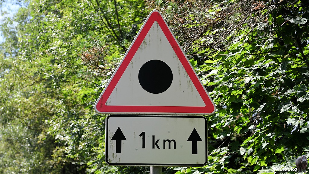 Das Bild zeigt ein dreieckiges Schild mit der Spitze nach oben mit rotem Rand. Darauf ist ein schwarzer Punkt auf weißem Grund. Darunter ist ein Zusatzschild mit der Aufschrift 1km. Die Schrift ist flankiert von zwei dicken Pfeilen nach oben.