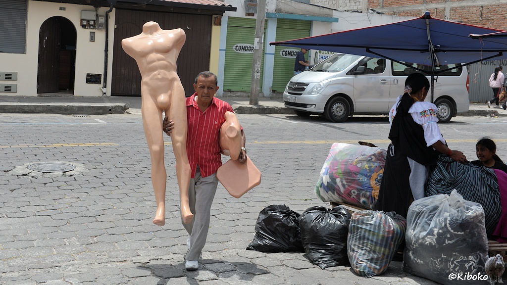 Das Bild zeigt einen Mann mit rotem Hemd und grauer Hose. Unter dem einen Arm hält er senkrecht eine männliche Schaufensterpuppe ohne Arme und ohne Kopf. Unter dem anderen Arm hat er eine weitere Schaufensterpuppe geklemmt.
