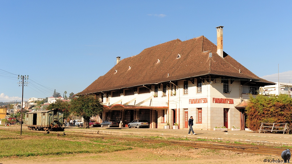Das Bild zeigt das zweigeschossige Bahnhofsgebäude mit einem über zwei Geschosse führendes hohes Walmdach. Unter dem ausladenden Vordach parken Autos im Schatten. Die Fenster und Türen sind rot geschtrichen. Auf dem Bahnsteig stehen zwei Bäume. Davor parkt ein grauer, vierachsiger gedeckter Güterwagen.