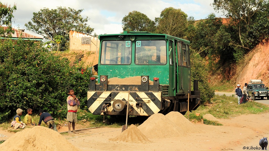 Das Bild zeigt eine grüne zweiachsige Motordraisine mit schwarz-gelben Warnanstrich and der Pufferbohle.  Neben der Strecke sind Sandhaufen udn Spaten. Vier kleine Kinder spielen neben und zwischen den Schienen.