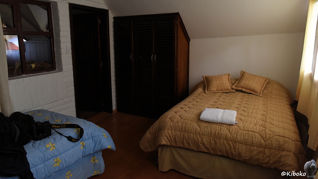 Das Bild zeigt einen Ausschnitt eines Zimmer mit einem braun bezogenen Doppelbett auf der rechten Seite und mit einem blau bezogenen Bett auf der linken Seite. Auf dem blauen Bett liegt ein Fotoapparat mit großem Objektiv. An der hinteren Wand steht ein Kleiderschrank, der an die Dachschräge angepasst wurde.