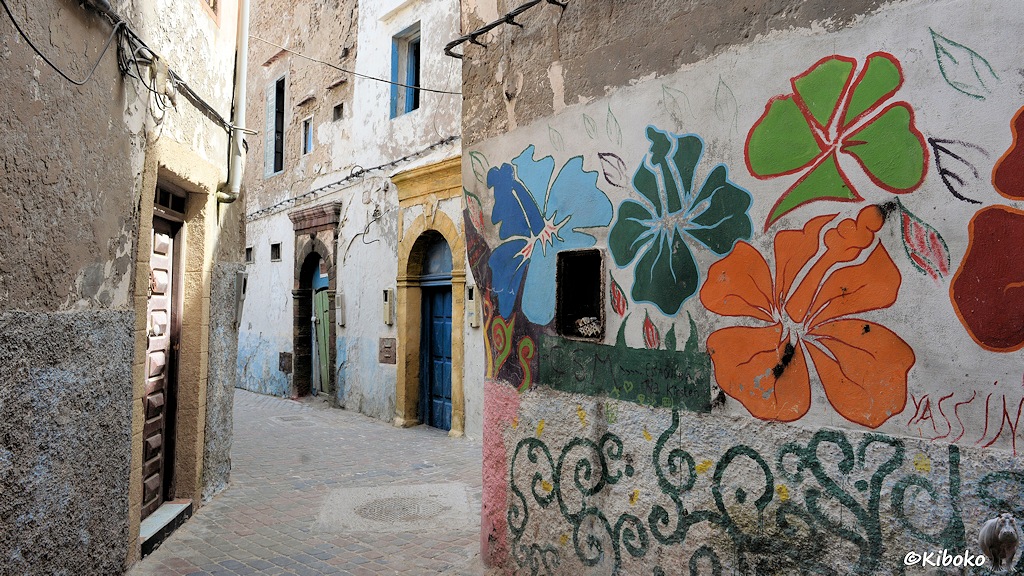 Das Bild zeigt eine enge Gasse, die einen Knick nach links macht. Die Hauswände sind schief. Der Putz blättert ab. Auf der rechten Seite wurden bunte Blumen an die Wand gemalt. Die Haustüren haben oft Rundbögen und sind windschief eingebaut.